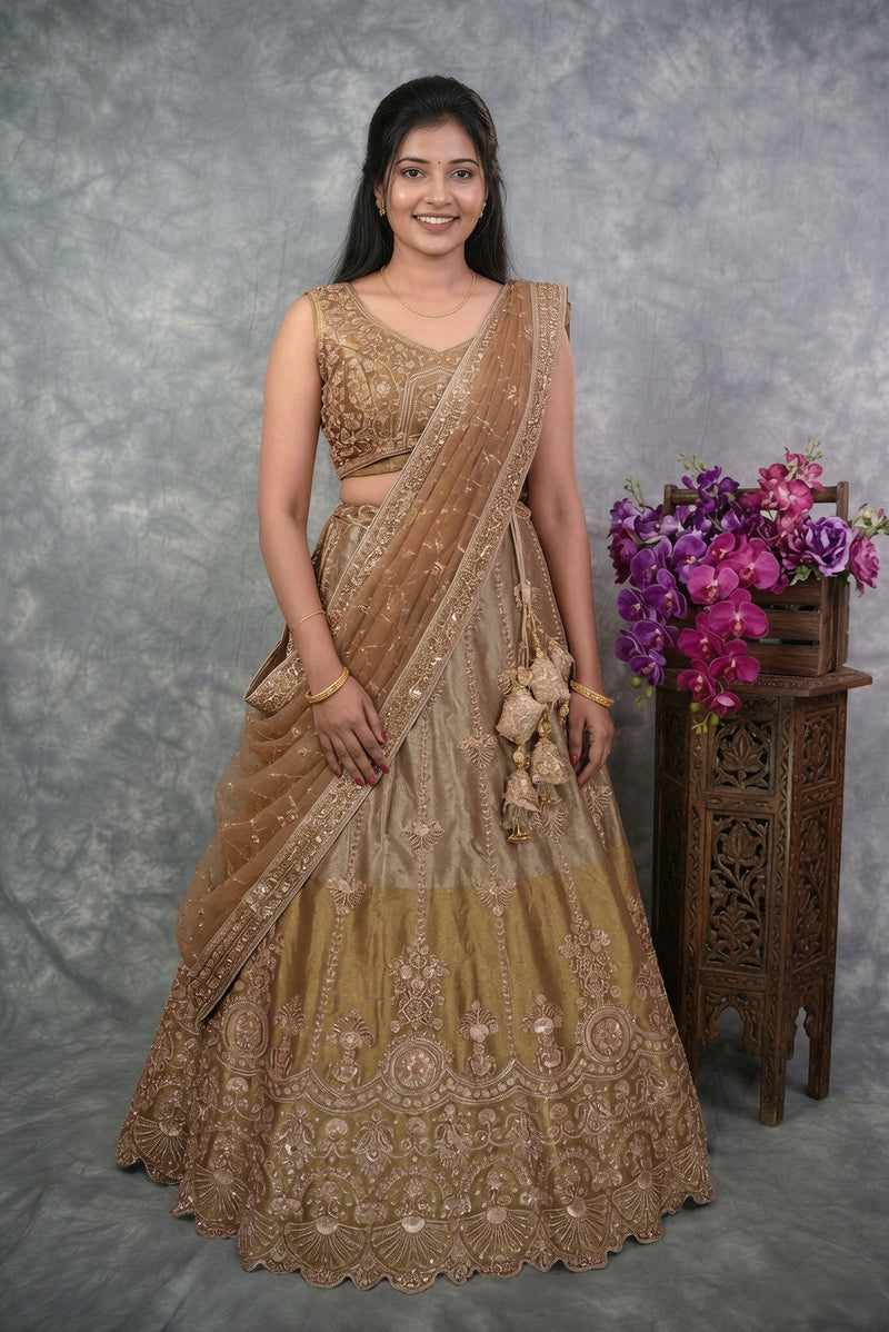 A woman modeling a traditional Indian lehenga choli set in a warm beige-gold color with intricate embroidery, standing against a textured gray backdrop next to a wooden stand with purple flowers.