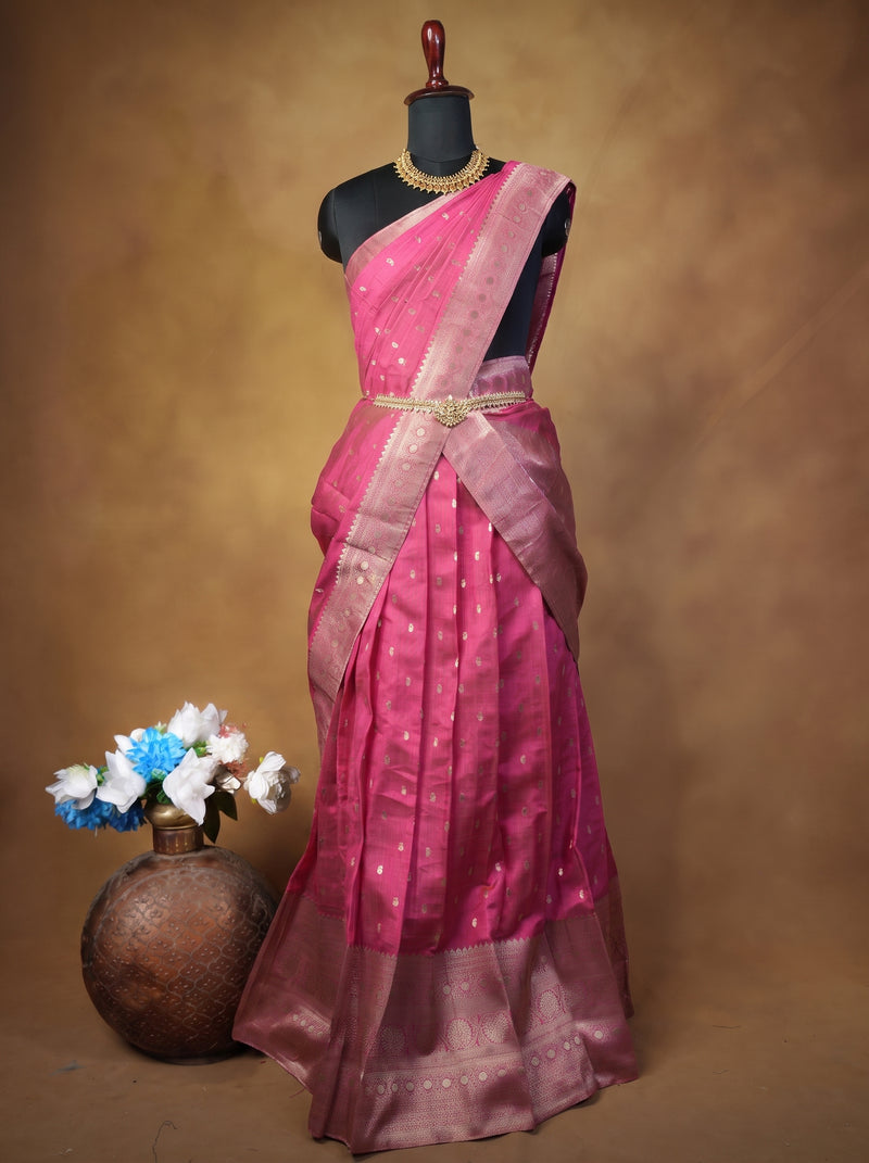 A pink silk saree with metallic gold zari embroidery and a matching pink border, displayed on a black mannequin with a decorative gold necklace. A rustic brown pot with blue and white flowers is placed on the left, set against a textured brown backdrop.