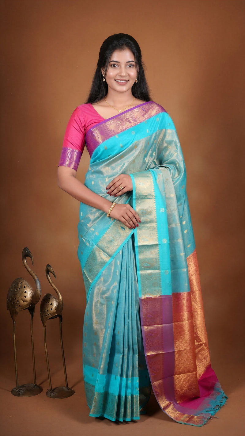 A woman modeling a traditional Indian saree with a turquoise blue body, magenta pink blouse, and gold zari borders, posed against a solid brown studio backdrop with decorative bird figurines at her side.