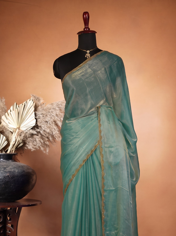 A sage green saree draped on a black mannequin torso, styled with a matching blouse piece. The saree features thin gold border accents.