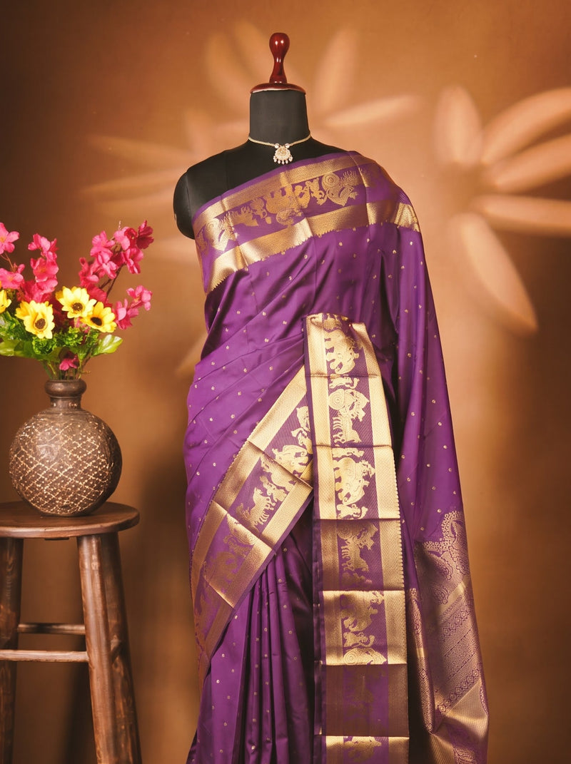 A purple silk saree with a wide gold zari border and motifs, displayed on a black mannequin. The mannequin is set against a warm brown background with decorative leaf shadows, and a wooden stool holding a vase of colorful flowers is on the left.