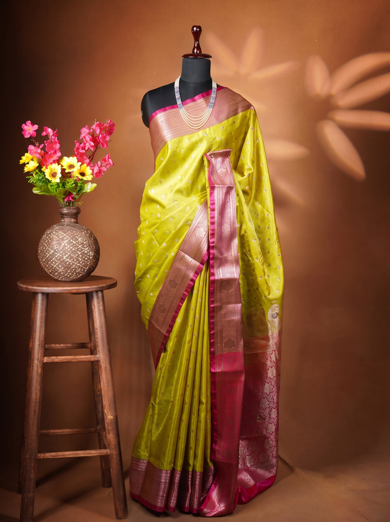 A yellow-green silk saree with a rich magenta-pink border and zari work, displayed on a black mannequin torso. The mannequin is styled with a pearl necklace and stands next to a small wooden stool holding a vase of pink and yellow flowers against a warm brown backdrop with leaf shadows.
