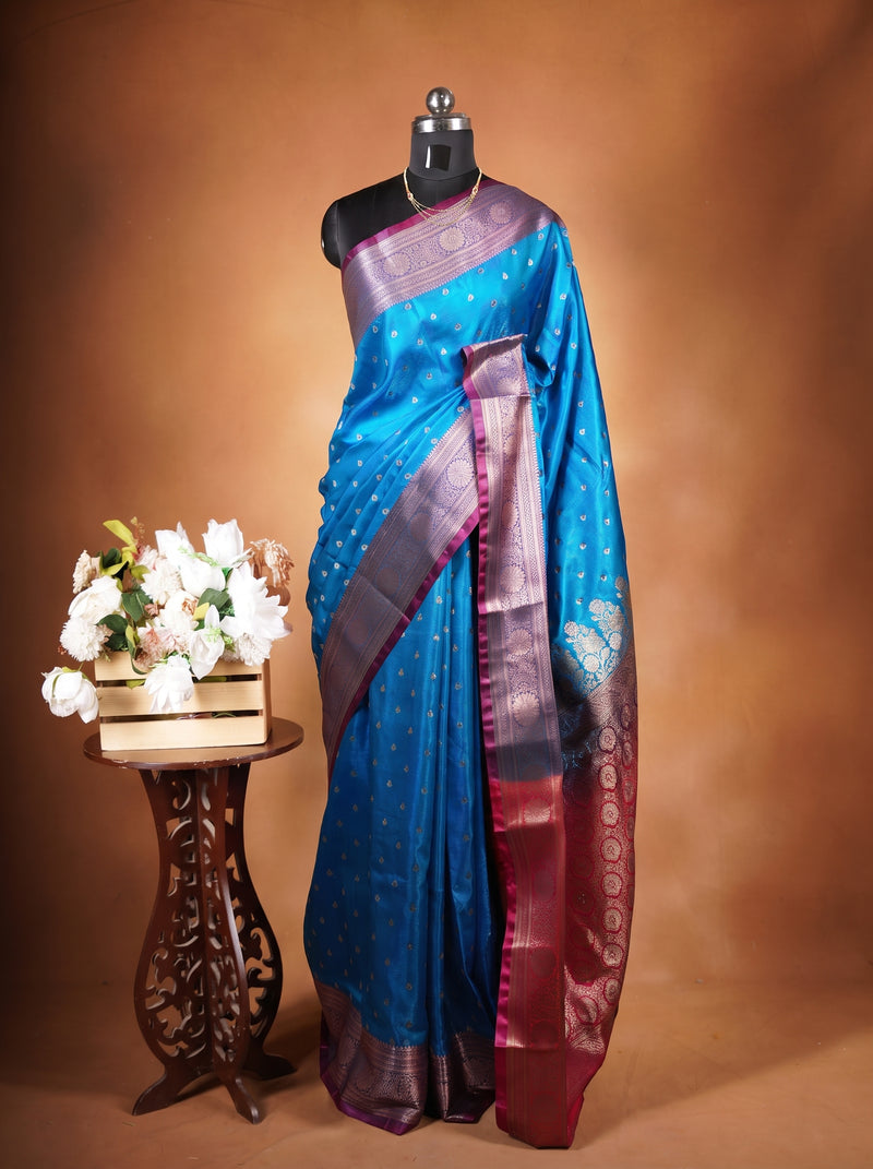 A blue and purple silk saree with gold zari motifs and a deep purple border, displayed on a black mannequin torso next to a small wooden table with white flowers, set against a warm brown studio background.