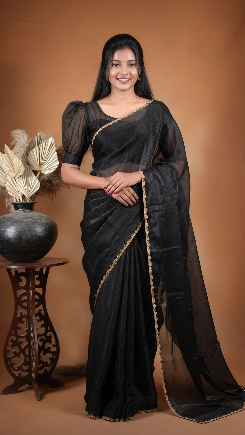 A woman modeling a black saree with gold border detailing, standing against a warm brown studio backdrop next to a decorative table with a vase.