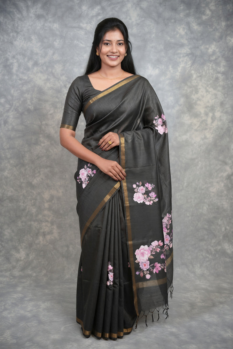 A woman modeling a dark gray saree with pink floral prints and gold borders, standing against a textured gray backdrop.