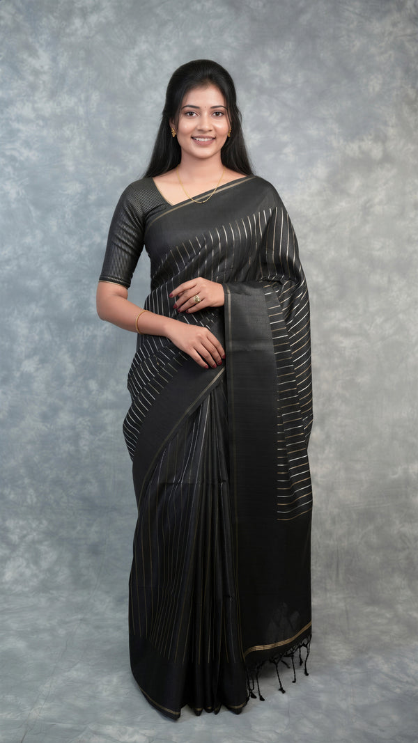 A woman modeling a black saree with subtle vertical metallic silver stripes, standing against a textured gray studio backdrop.