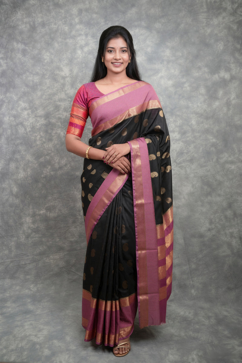 A woman modeling a black and pink silk saree with gold zari motifs and borders, standing against a textured gray studio backdrop.