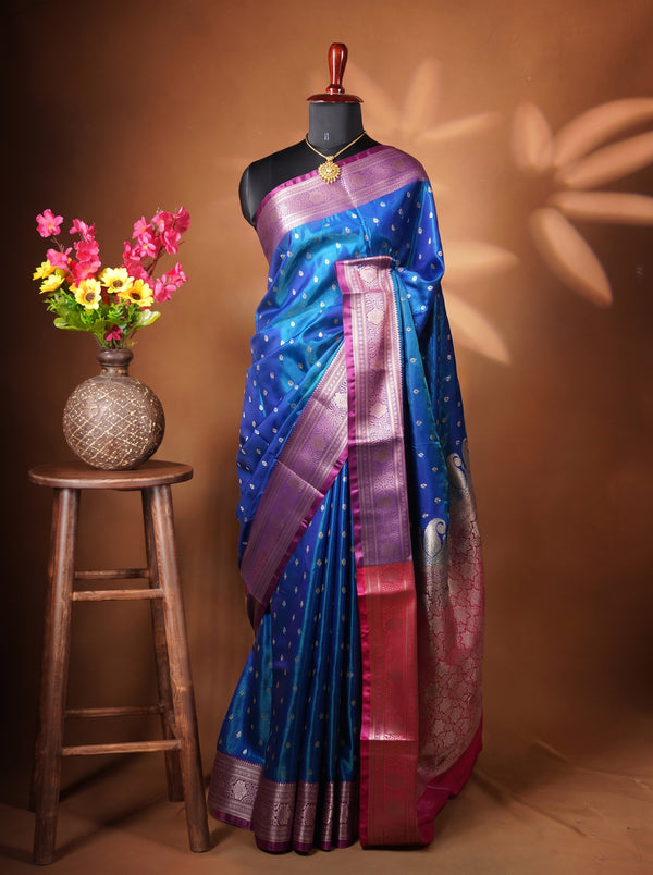 A blue and purple silk saree with gold zari embroidery and a red border, displayed on a black mannequin torso. The mannequin is set against a warm brown backdrop with leaf shadows, next to a wooden stool holding a vase of pink and yellow flowers.
