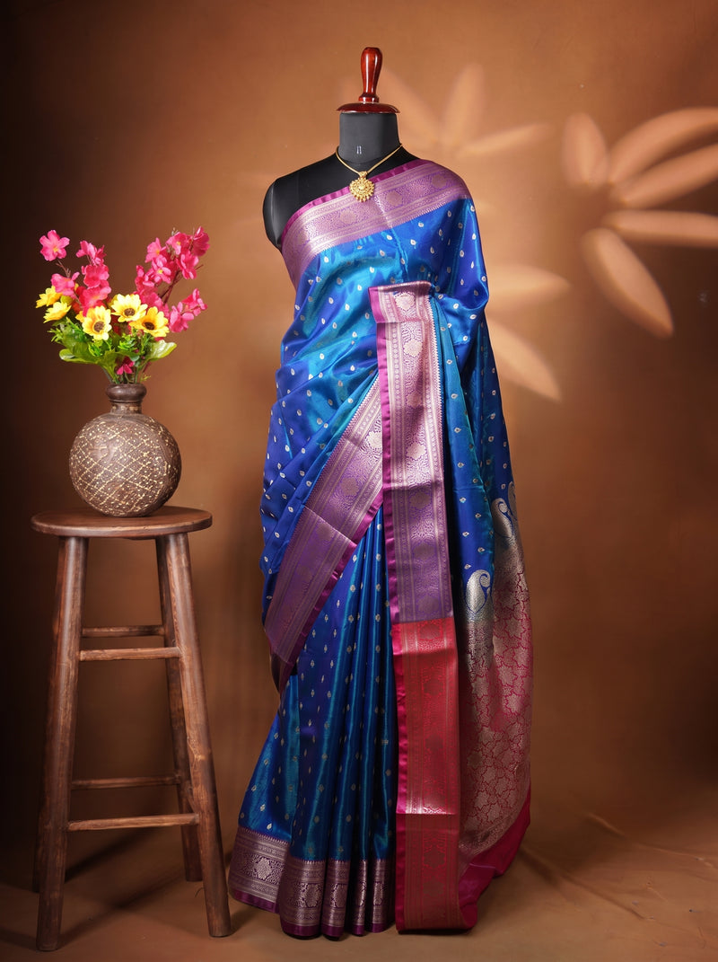A blue and purple silk saree with gold zari embroidery and a red border, displayed on a black mannequin torso. The mannequin is set against a warm brown backdrop with leaf shadows, next to a wooden stool holding a vase of pink and yellow flowers.