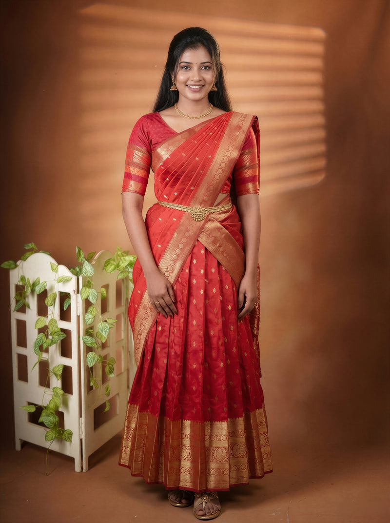 A woman wearing a red and gold traditional Indian saree stands against a warm brown studio backdrop with a decorative white screen and green vines on the left.