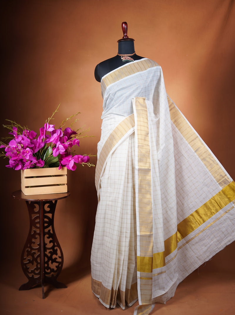 A white saree with gold borders displayed on a black mannequin torso, set against a warm brown studio backdrop. A small wooden stand holding bright pink flowers is positioned to the left.
