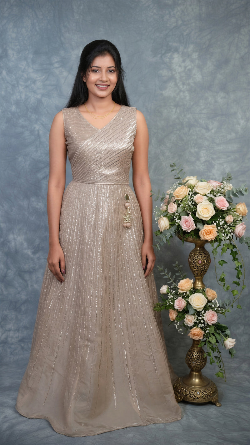 A woman wearing a sleeveless, floor-length champagne beige gown with shimmering sequin details, standing against a mottled gray studio backdrop next to a decorative floral arrangement on a pedestal.