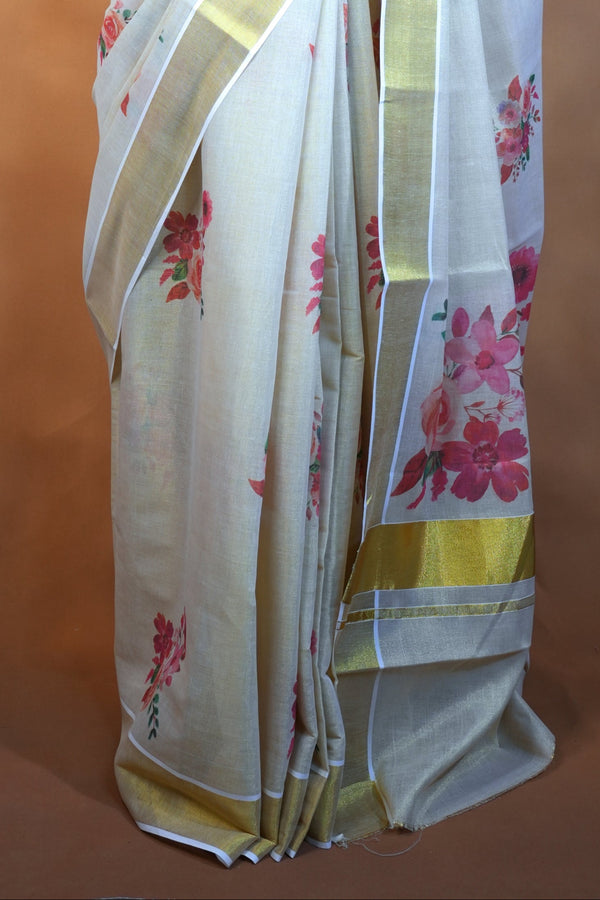 A white silk saree with pink floral prints and gold borders, draped on a black mannequin torso against a warm brown textured backdrop.