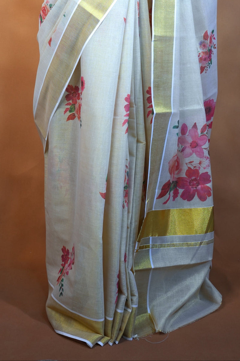 A white silk saree with pink floral prints and gold borders, draped on a black mannequin torso against a warm brown textured backdrop.