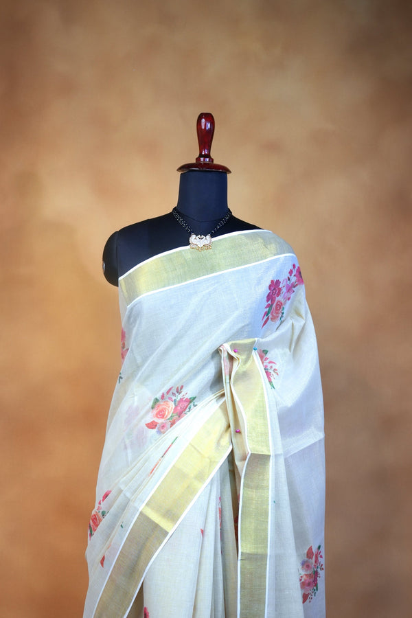 A white silk saree with pink floral prints and gold borders, draped on a black mannequin torso against a warm brown textured backdrop.