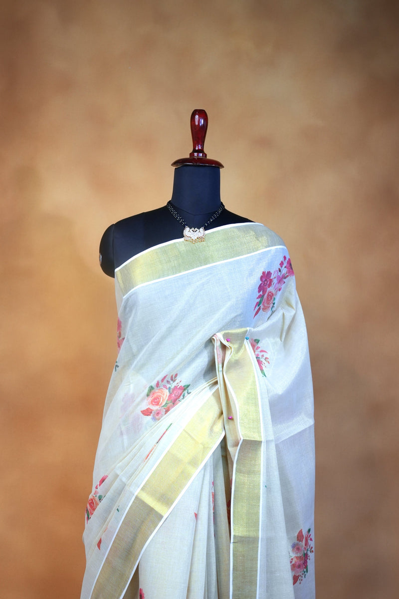 A white silk saree with pink floral prints and gold borders, draped on a black mannequin torso against a warm brown textured backdrop.