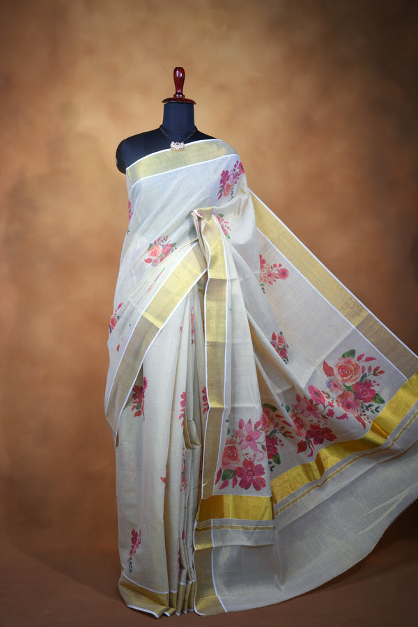 A white silk saree with pink floral prints and gold borders, draped on a black mannequin torso against a warm brown studio backdrop.