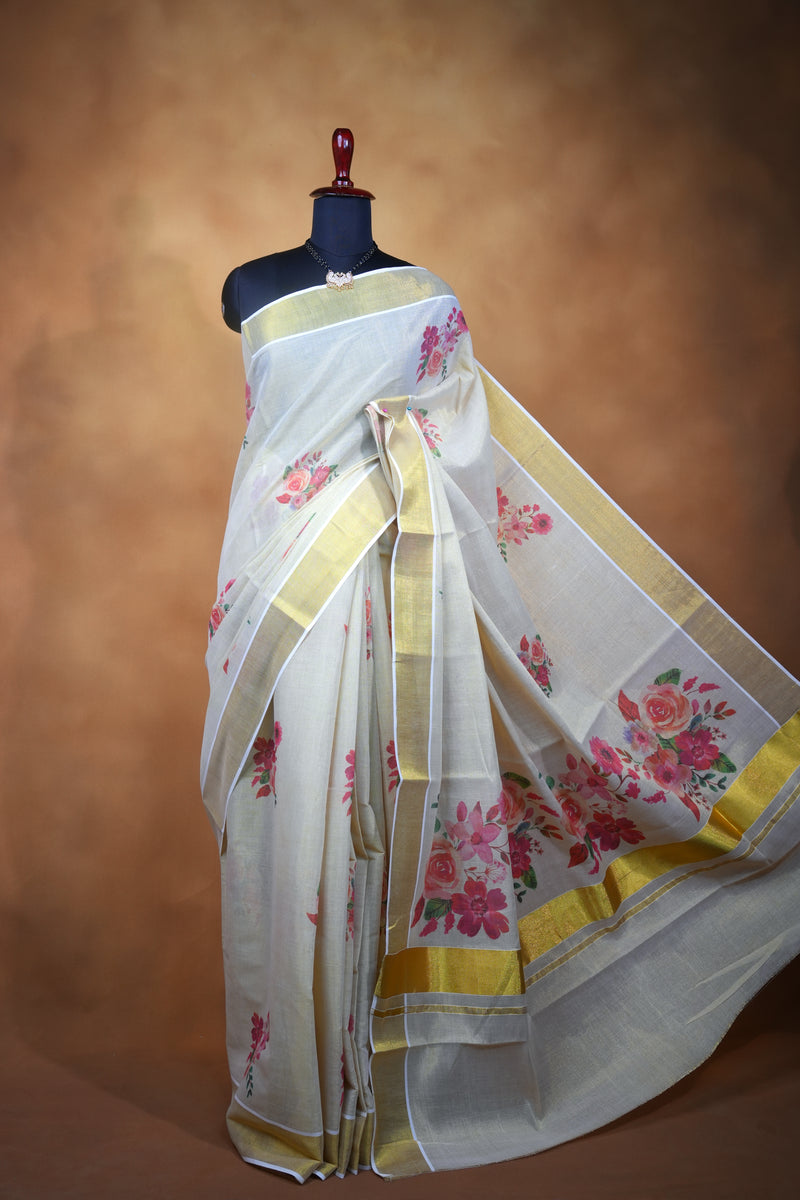 A white silk saree with pink floral prints and gold borders, draped on a black mannequin torso against a warm brown studio backdrop.