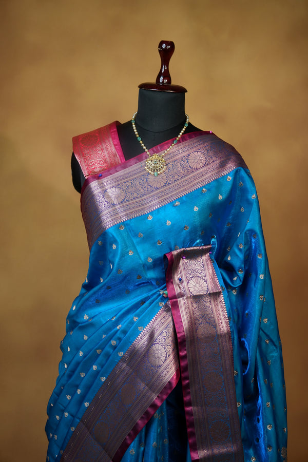 A blue silk saree with a metallic gold zari border and pinkish-purple pallu, draped elegantly on a black mannequin torso against a warm brown background.