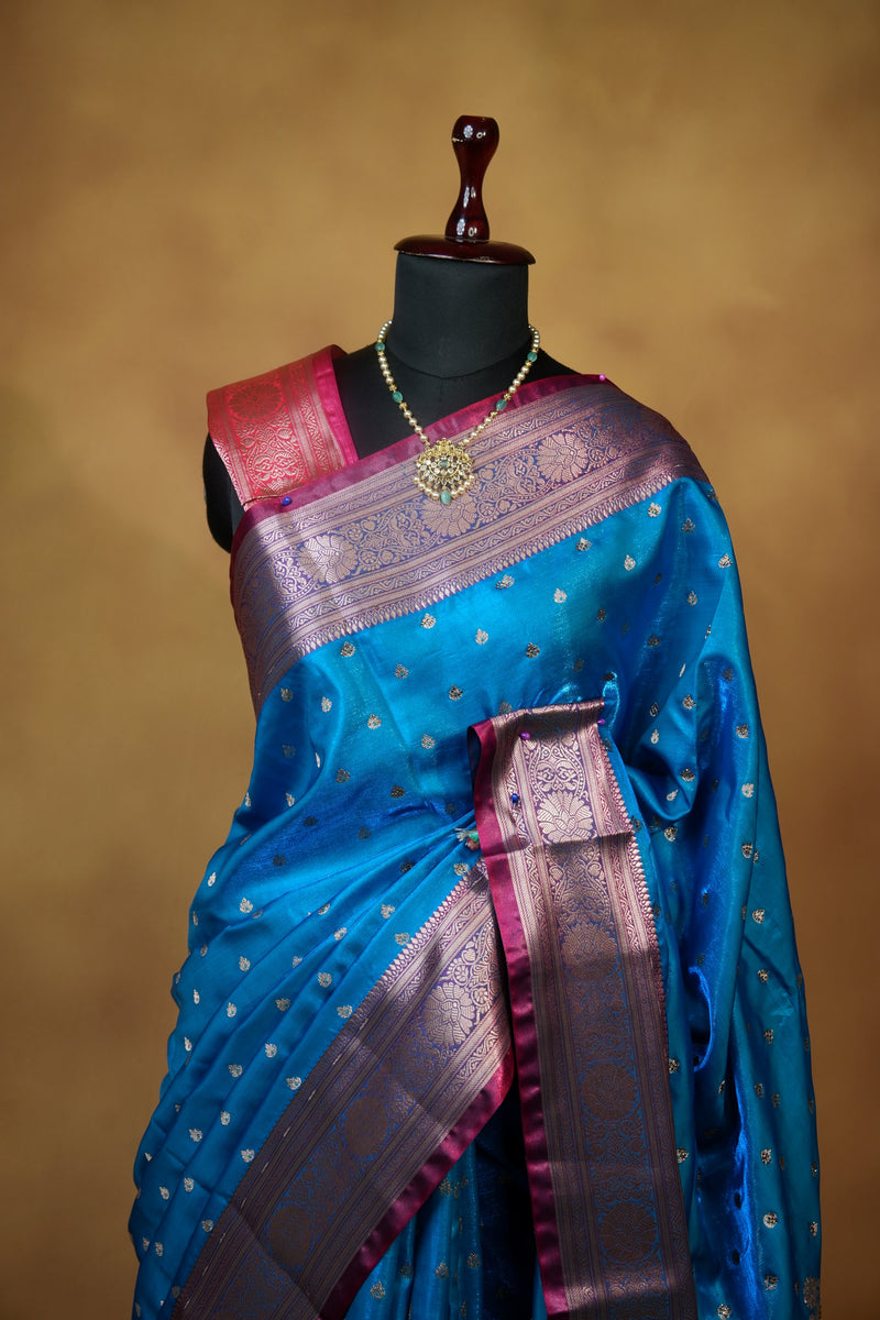 A blue silk saree with a metallic gold zari border and pinkish-purple pallu, draped elegantly on a black mannequin torso against a warm brown background.