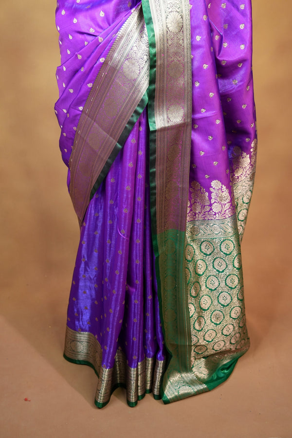 A folded purple silk saree with a green border, Gold zari motifs, and a metallic silver pallu, displayed on a neutral beige surface.