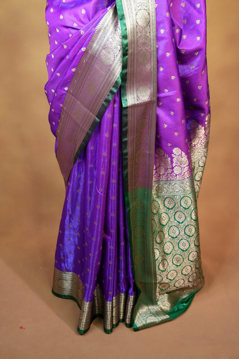 A folded purple silk saree with a green border, Gold zari motifs, and a metallic silver pallu, displayed on a neutral beige surface.
