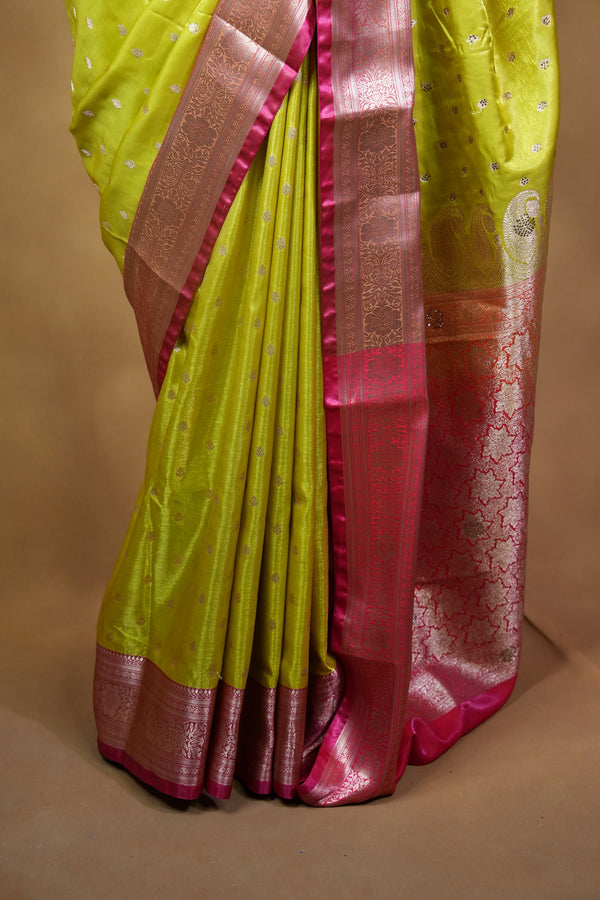 A folded yellow-green silk saree with a metallic pink border and silver zari motifs, displayed inside a cardboard box.