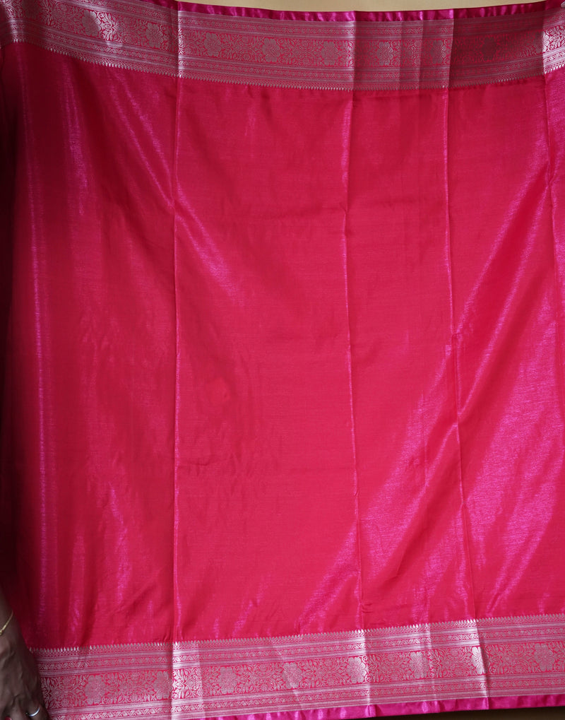 A close-up of a bright pink fabric, likely a saree, featuring a shimmering pink border with metallic gold and silver zari patterns.