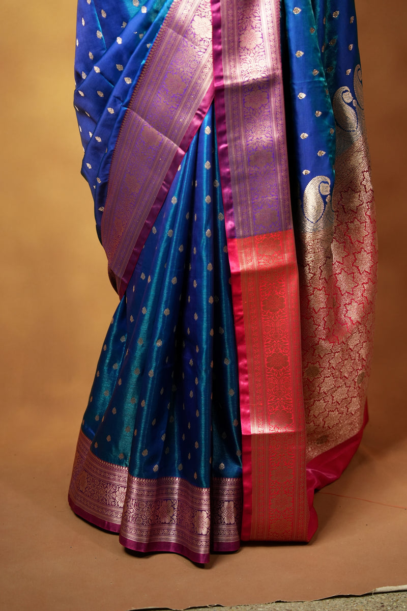 A close-up of a blue silk saree with gold zari motifs, a pink and purple border, and a red pallu, draped against a solid tan background.