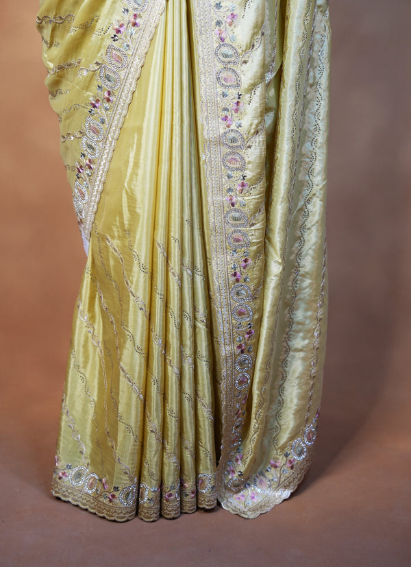 A close-up of a draped yellow-gold fabric with ornate metallic embroidery and small floral accents, shown against a plain brown studio background.