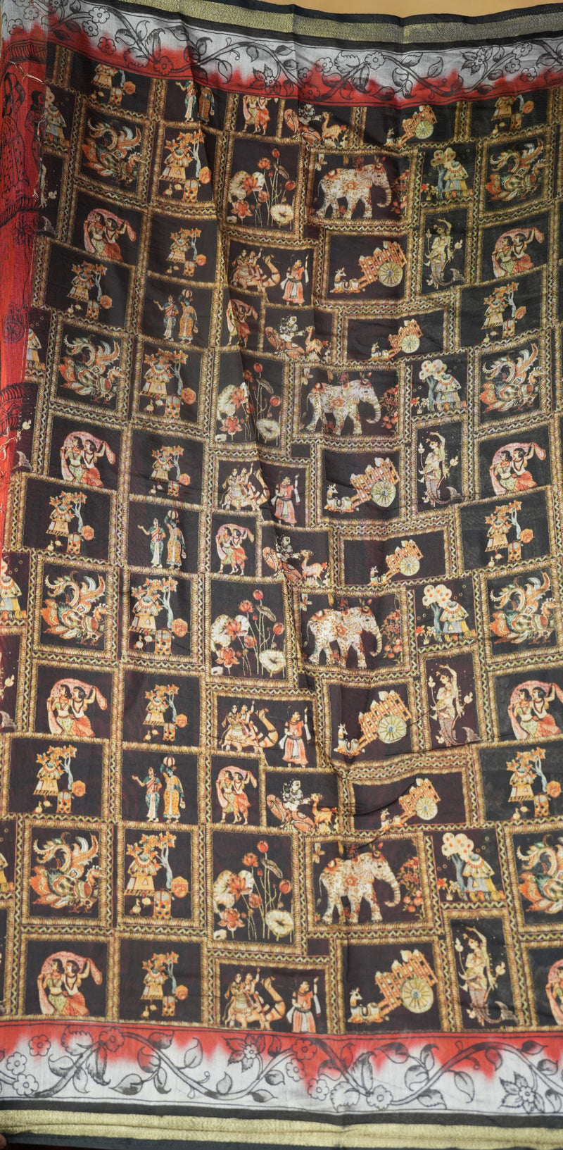 A black fabric textile featuring a grid of small illustrated panels with figures, elephants, and decorative motifs in gold, red, beige, and white, plus a patterned border at the top and bottom.