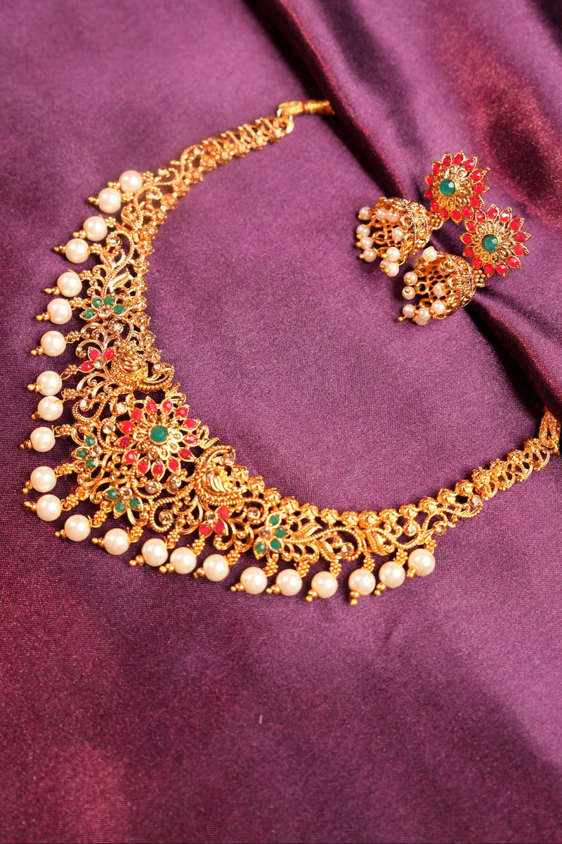 An ornate gold-toned traditional Indian tiara crown decorated with white pearls, red, green, and gold stones, displayed with matching earrings on a draped purple satin fabric.