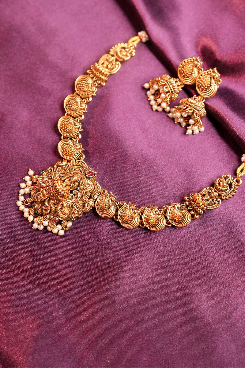 An ornate gold-toned traditional necklace with matching earrings, displayed on draped purple satin fabric.