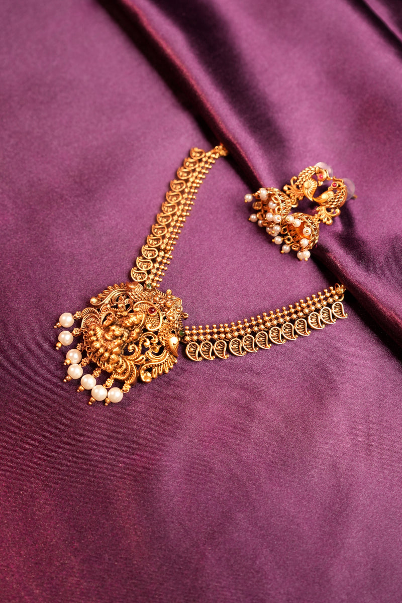 An ornate gold-toned traditional Indian hair ornament (jhumka) with pearl embellishments, displayed on a draped purple satin fabric.