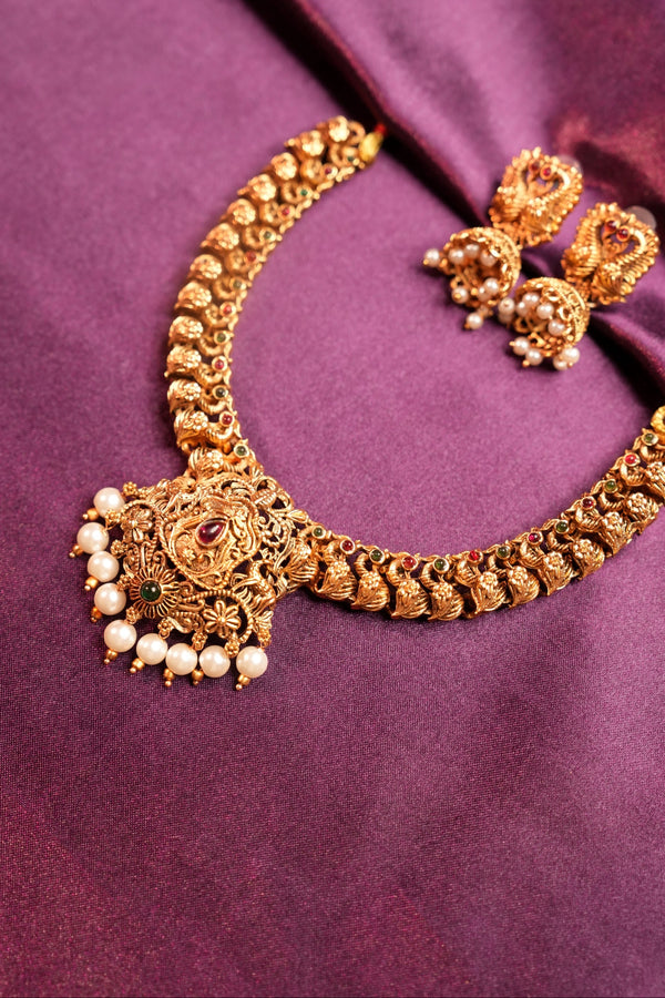 An ornate gold-toned traditional curved necklace with pearl accents and a matching pair of earrings, displayed on a draped purple fabric background.