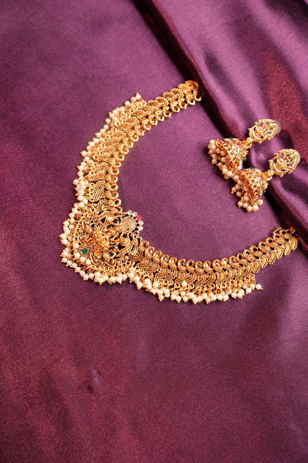 An ornate gold-toned traditional jewelry set featuring a heavily embellished curved necklace and matching earrings, displayed on a draped deep purple satin fabric.