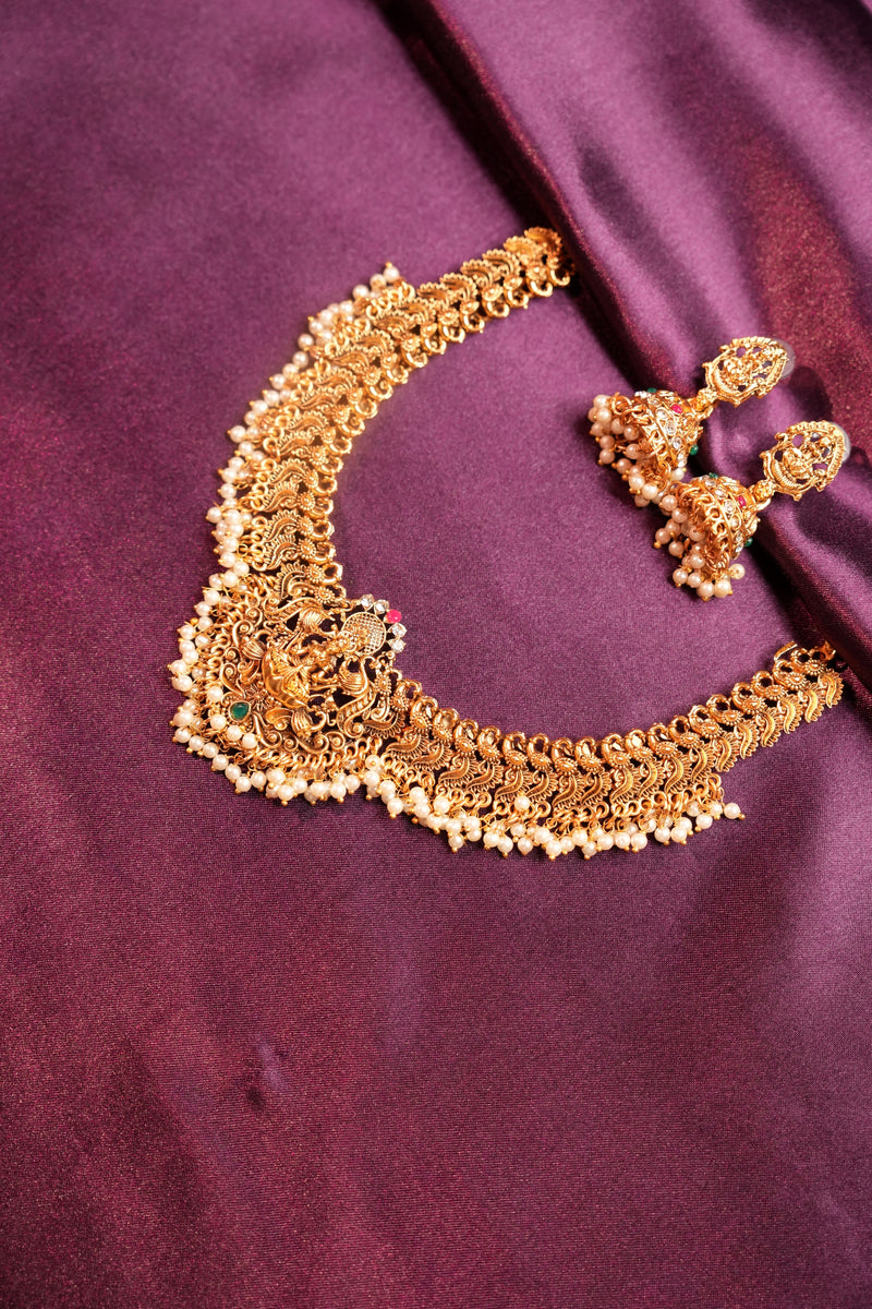 An ornate gold-toned traditional jewelry set featuring a heavily embellished curved necklace and matching earrings, displayed on a draped deep purple satin fabric.