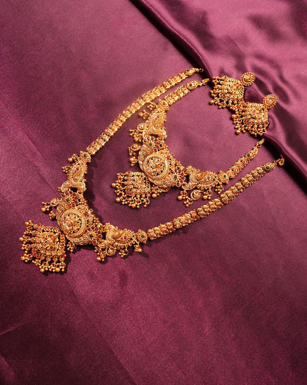 An ornate gold-toned traditional jewelry set with a necklace, earrings, and matching bracelet arranged on a rich magenta satin fabric background.