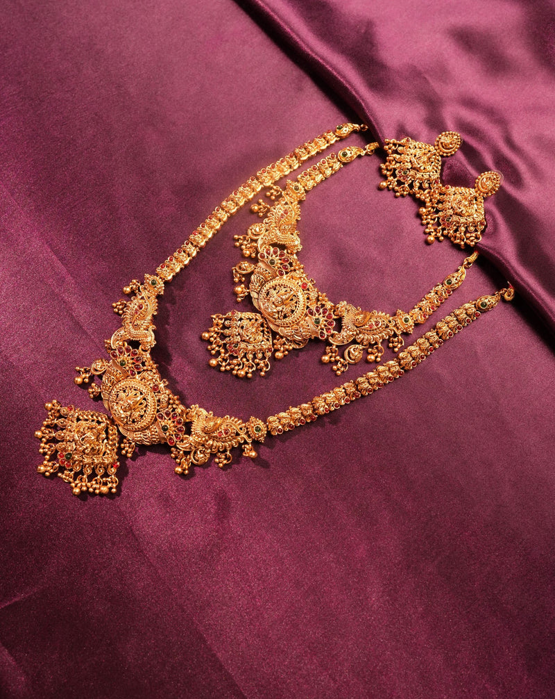 An ornate gold-toned traditional jewelry set with a necklace, earrings, and matching bracelet arranged on a rich magenta satin fabric background.