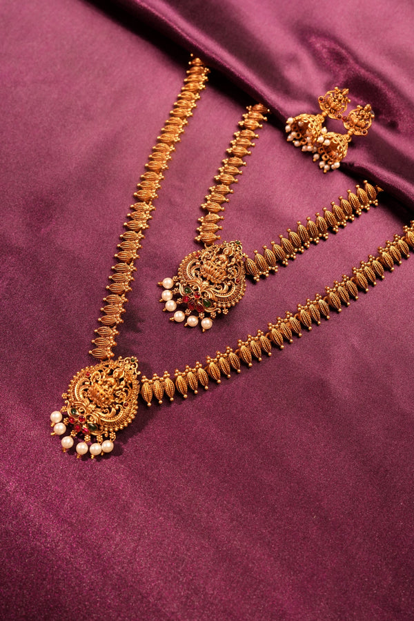 An ornate gold-toned traditional necklace set with matching earrings and a choker, decorated with intricate filigree, small white pearl-like beads, and red stone accents, arranged on a draped purple satin fabric.