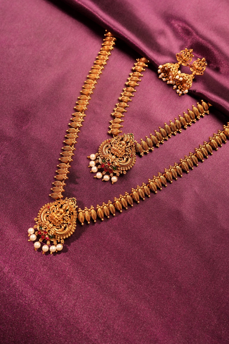 An ornate gold-toned traditional necklace set with matching earrings and a choker, decorated with intricate filigree, small white pearl-like beads, and red stone accents, arranged on a draped purple satin fabric.