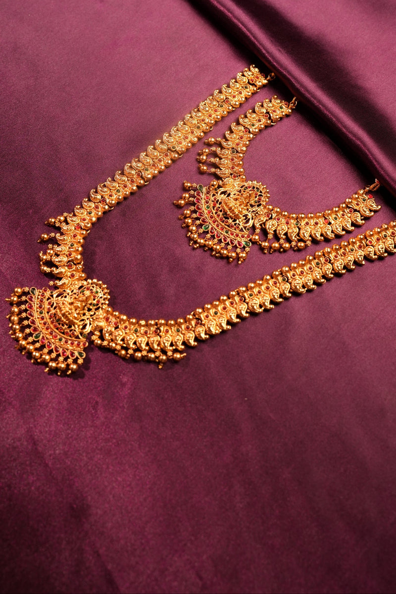 A pair of ornate gold-toned traditional jewelry pieces, likely Indian Kundan necklaces or maang tikka sets, displayed on a draped deep purple satin fabric.