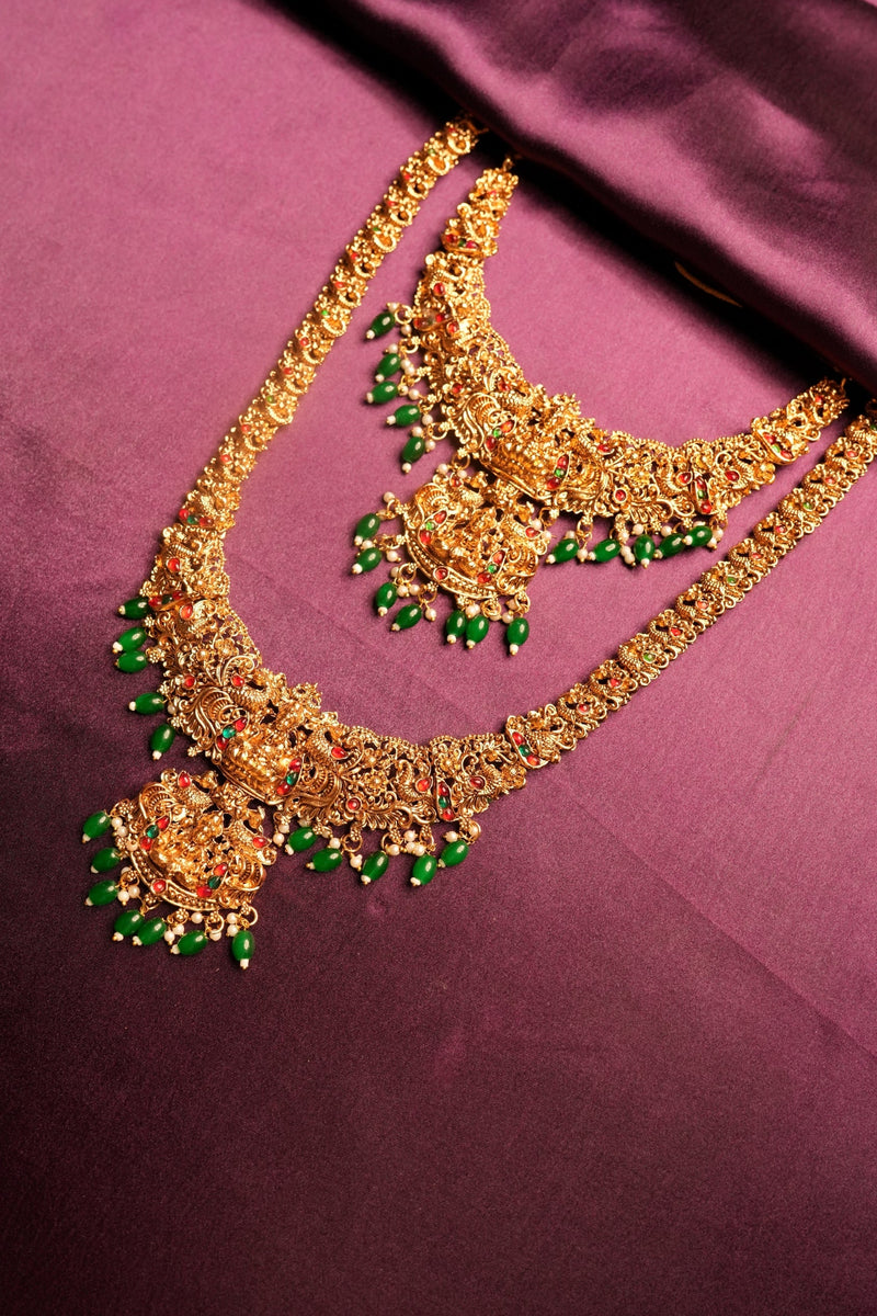 Gold-toned traditional Indian jewelry set featuring a large arched necklace and a matching curved pendant, both adorned with green stone accents, displayed on a draped magenta satin fabric.