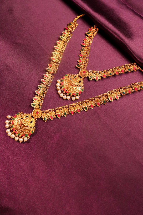 An ornate gold-toned traditional necklace set with red and green stone accents and white bead fringe, displayed on a draped deep purple fabric background.