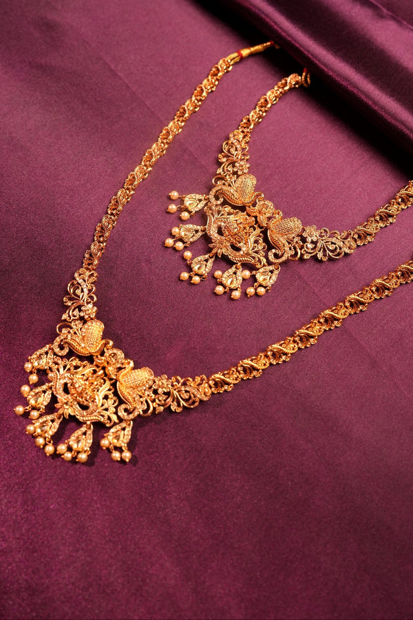 An ornate gold-toned traditional necklace with intricate filigree, floral motifs, and dangling bead details, displayed on draped deep purple fabric.