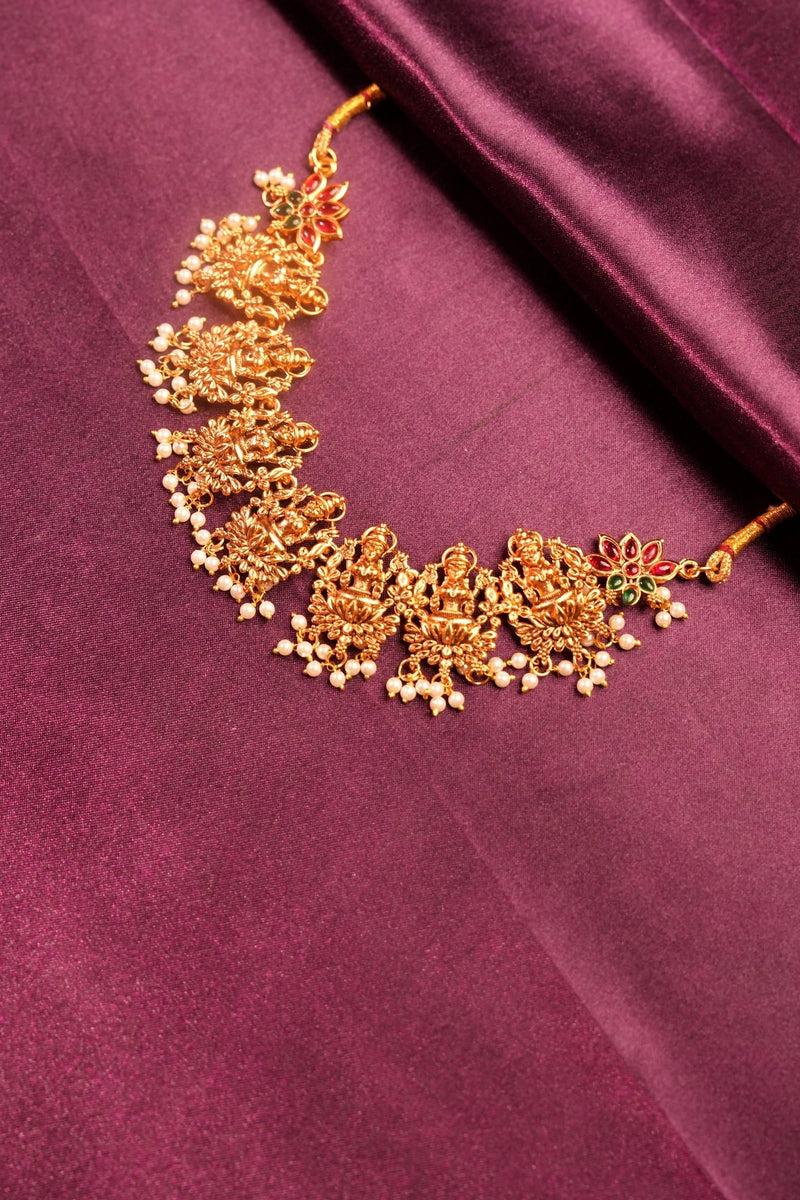 Gold-toned traditional Indian bridal hair ornament (jhumka) with small white floral embellishments and red stone accents, displayed on a draped purple satin fabric.