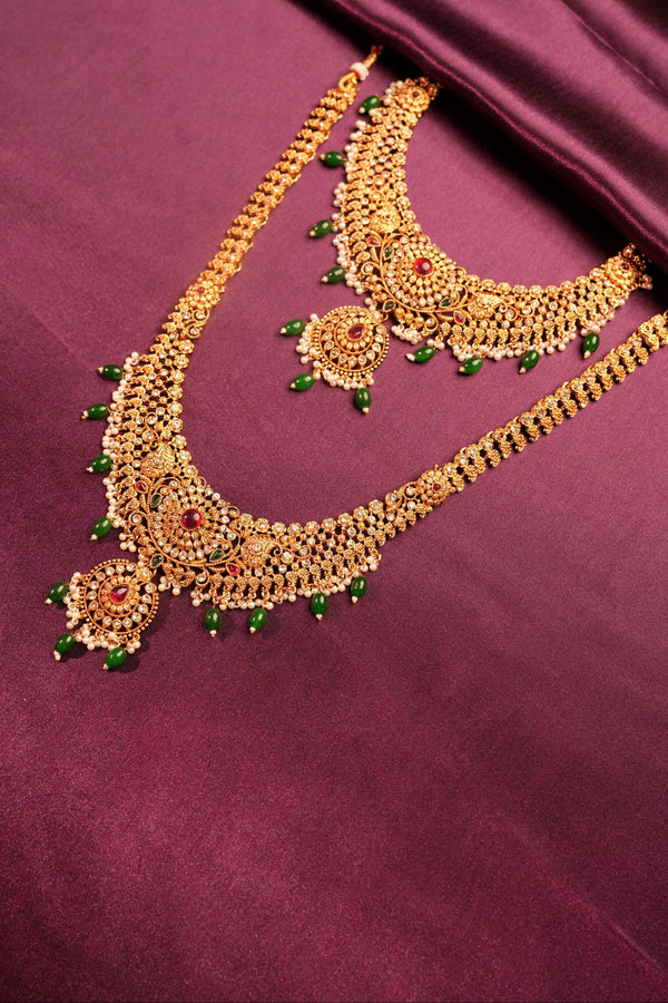 An ornate gold-toned traditional necklace set with green bead accents and red stone details, arranged on a draped deep purple fabric background.