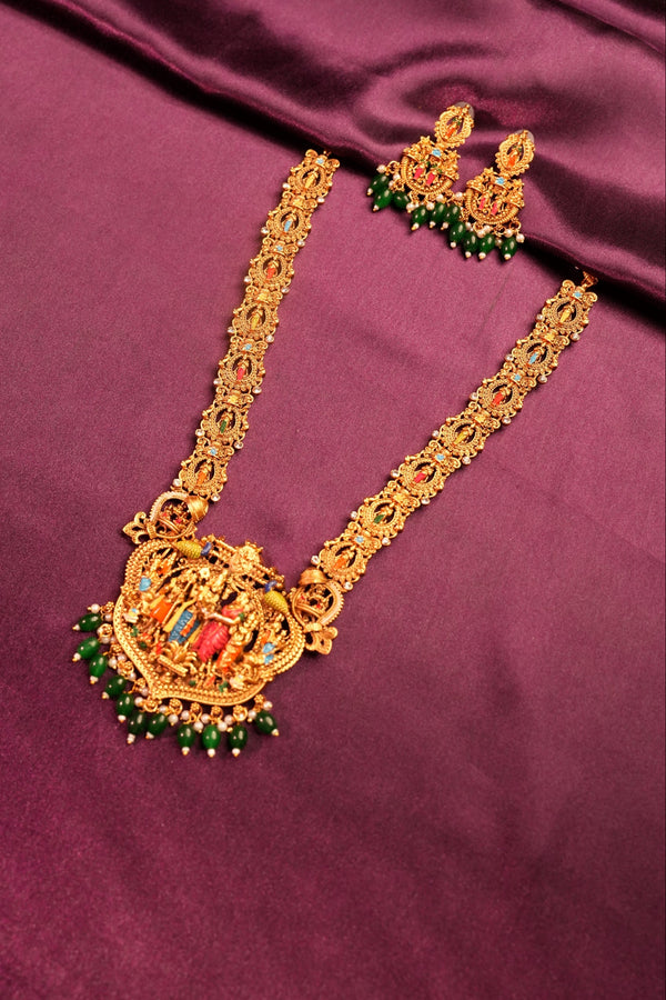 An ornate gold-toned traditional Indian jewelry piece with green and red gemstone accents, displayed on a draped purple satin fabric.