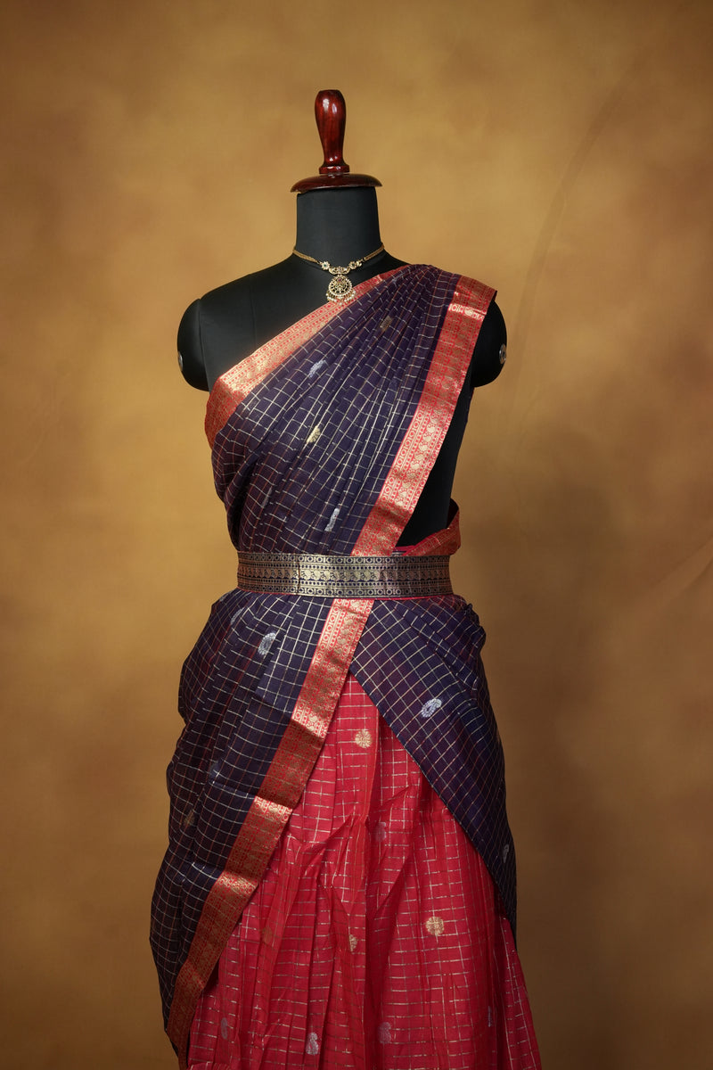A folded traditional garment draped on a black mannequin torso against a warm brown studio background. The fabric shows a red body with a dark navy-blue grid pattern and metallic copper-gold borders.