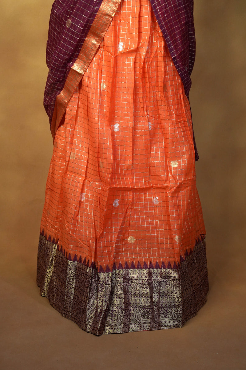 An orange silk saree with a purple border and a matching purple checkered dupatta draped over the shoulders, set against a plain brown background.